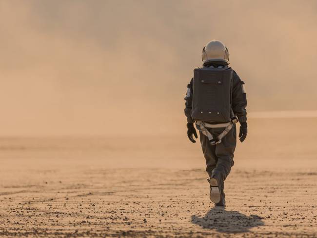 Foto de un astronauta con traje espacial y casco en Marte alejándose de la cámara hacia una tormenta de polvo a lo lejos