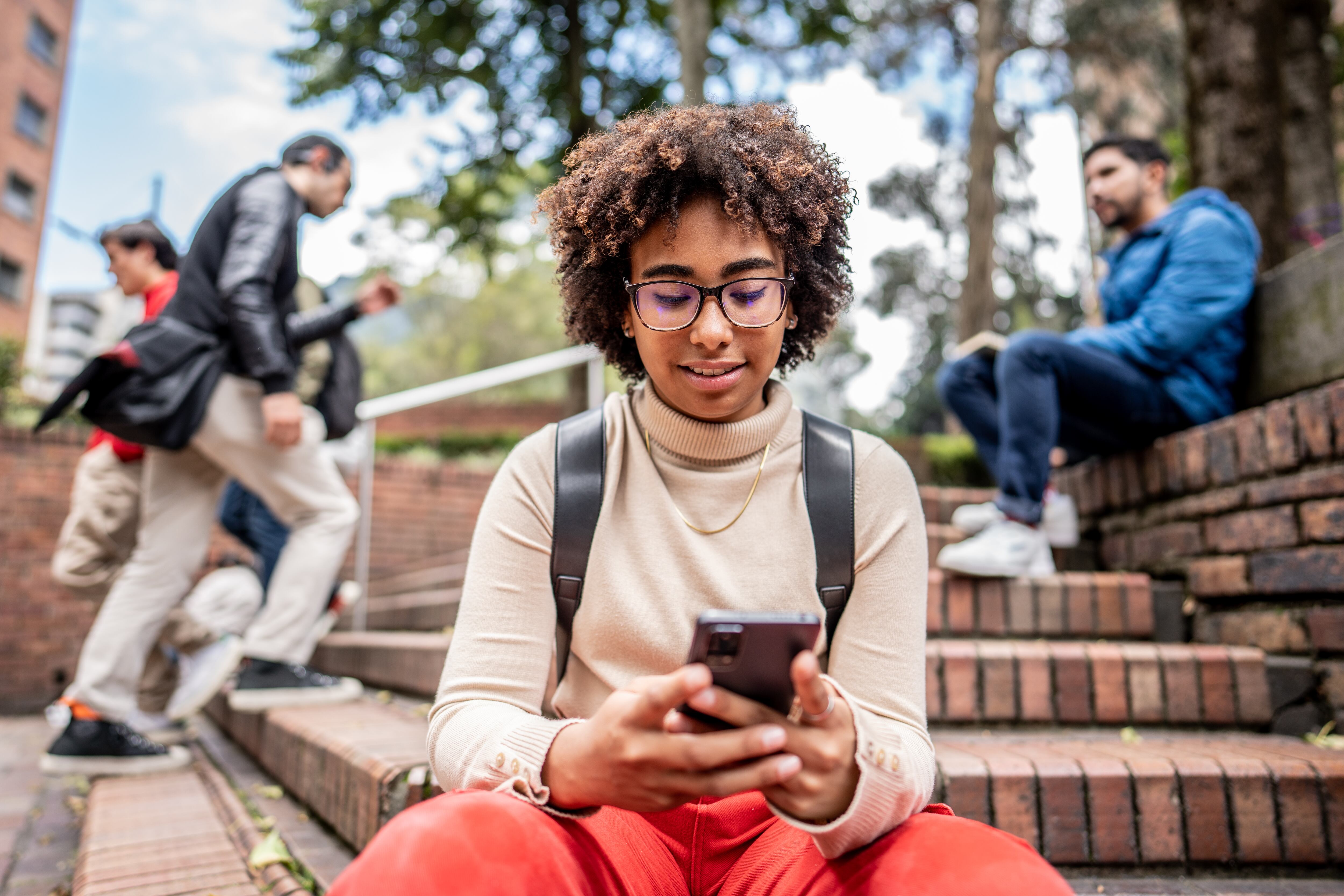 Estudiante universitaria utilizando su celular mientras está sentada en las escaleras del campus (Foto vía Getty Images)
