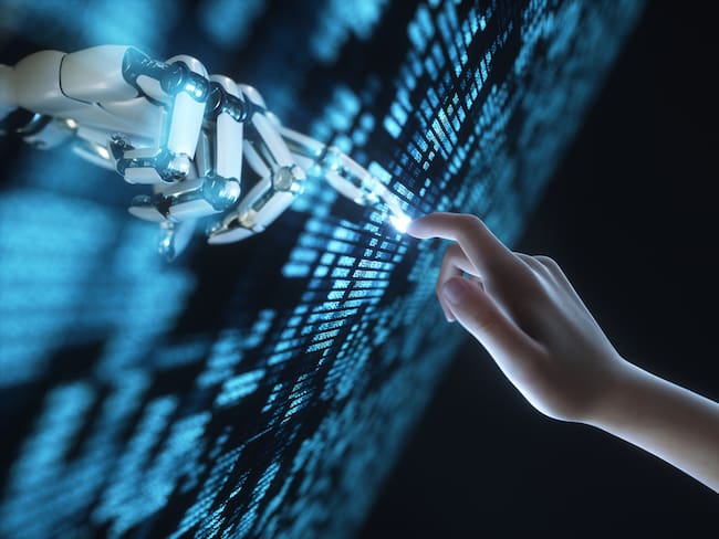 Imagen generada digitalmente de la mano del robot tocando la mano del ser humano a través de un panel digital transparente con números sobre fondo negro / Getty Images