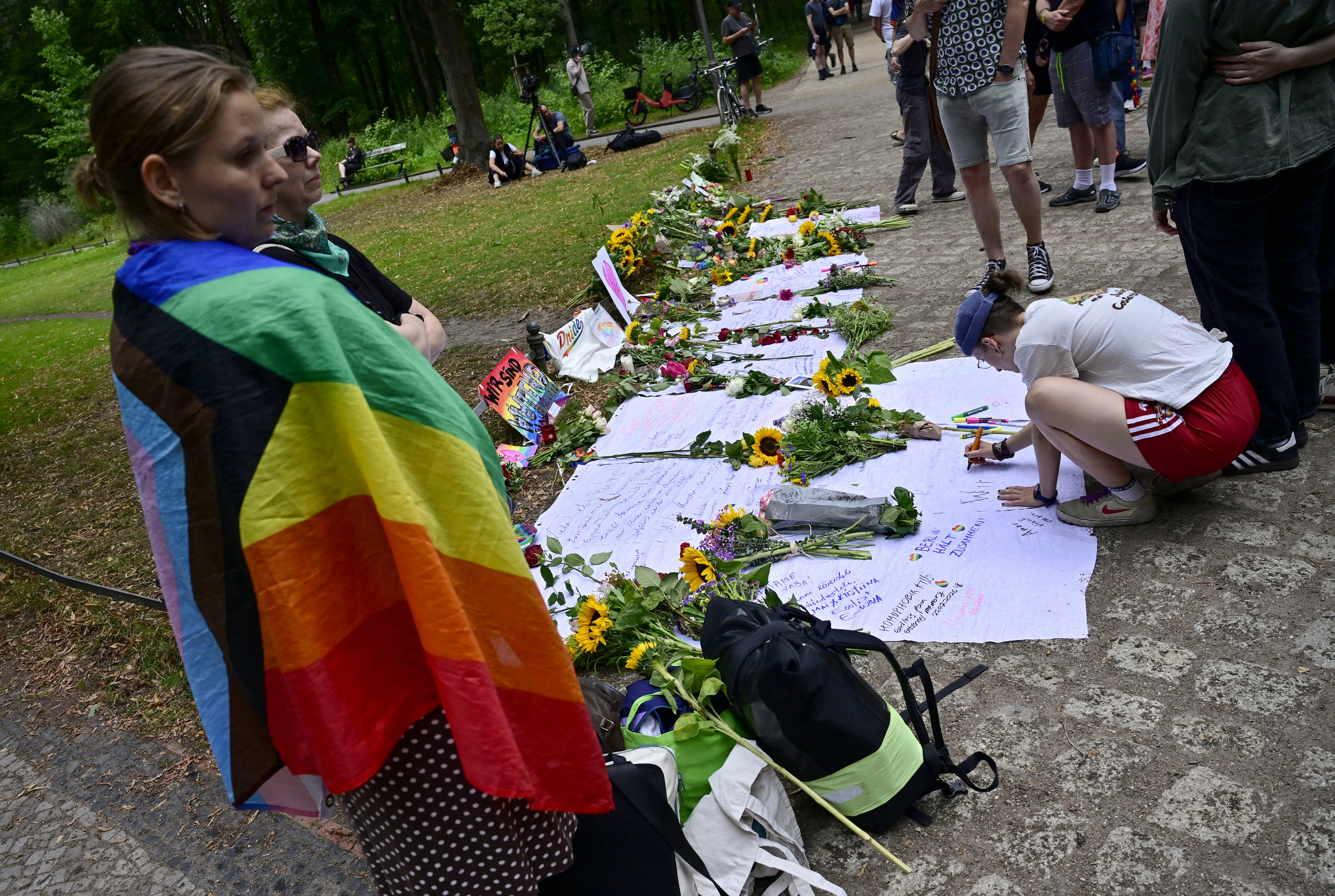 Un doliente envuelto en una bandera arcoíris observa cómo otros dejan un mensaje junto a flores en un monumento improvisado para las víctimas el 26 de julio de 2026 cerca del lugar en el parque Tiergarten de Berlín, donde la noche anterior un automóvil atropelló a varias personas, matando a una e hiriendo a 16 en las inmediaciones del desfile del Día de Christopher Street (CSD). La policía alemana buscó el 26 de julio de 2026 a un sospechoso presuntamente vinculado a círculos islamistas después de que un automóvil embistiera a una multitud cerca de las celebraciones del Orgullo Gay de Berlín, matando a una persona e hiriendo a otras 16. El incidente empañó lo que había sido un día de celebración para cientos de miles de asistentes al Día de Christopher Street, uno de los eventos del Orgullo más grandes de Europa, y provocó que los asistentes huyeran de un parque céntrico de Berlín mientras las sirenas resonaban en la noche. (Foto de John MACDOUGALL / AFP)