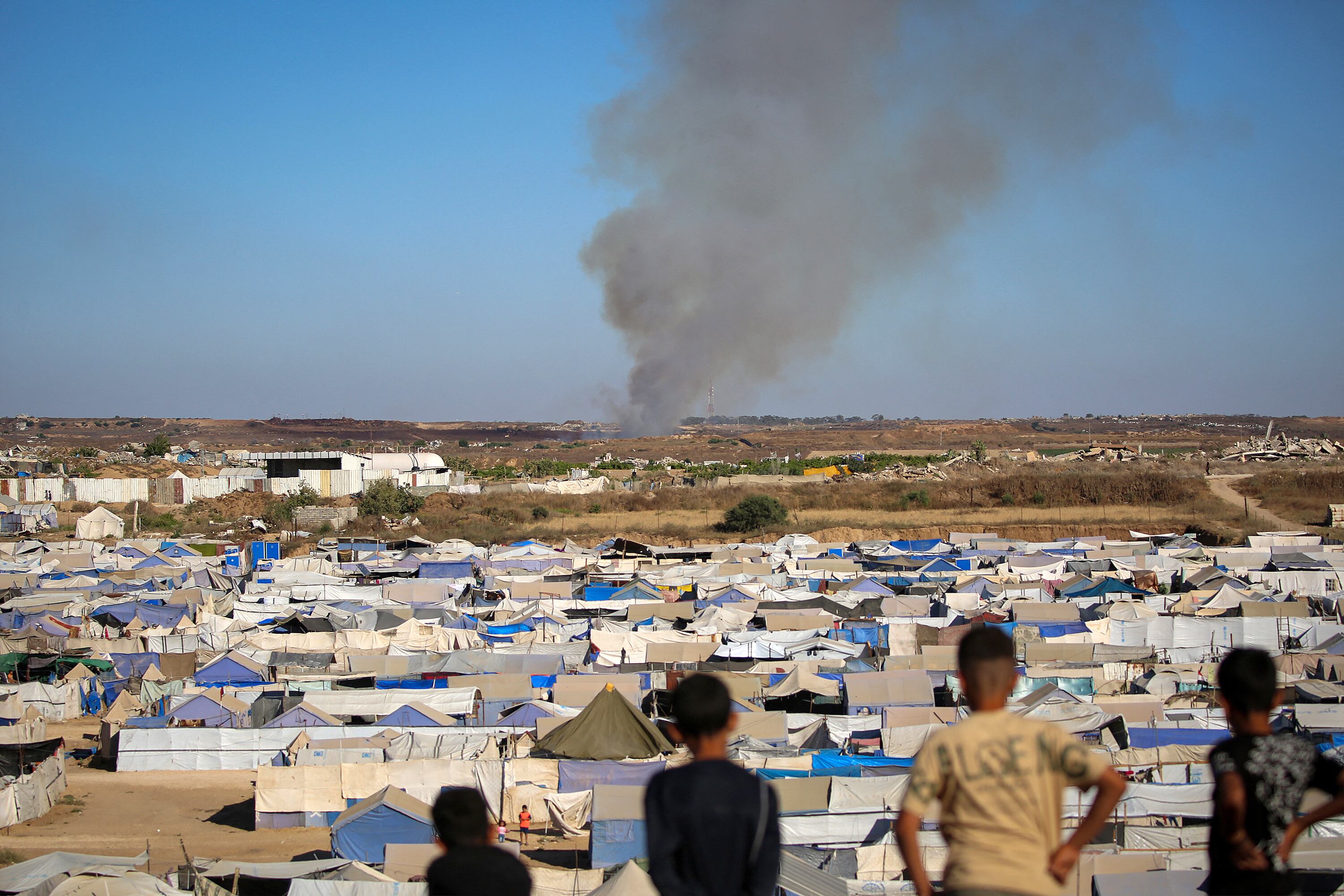 Children watch from atop a promontory overlooking shelters for people displaced by war, as smoke billows in the background near the Nuseirat camp for Palestinian refugees in the central Gaza Strip on June 19, 2026. Palestinian Islamist movement Hamas on Monday hailed a deal between the United States and Iran, expressing hope it would help put an end to violence in the Gaza Strip. Israel and Hamas trade near-daily accusations of truce violations and the Gaza Strip remains gripped by bloodshed as progress on permanently ending the war remains stalled. (Photo by Eyad Baba / AFP)