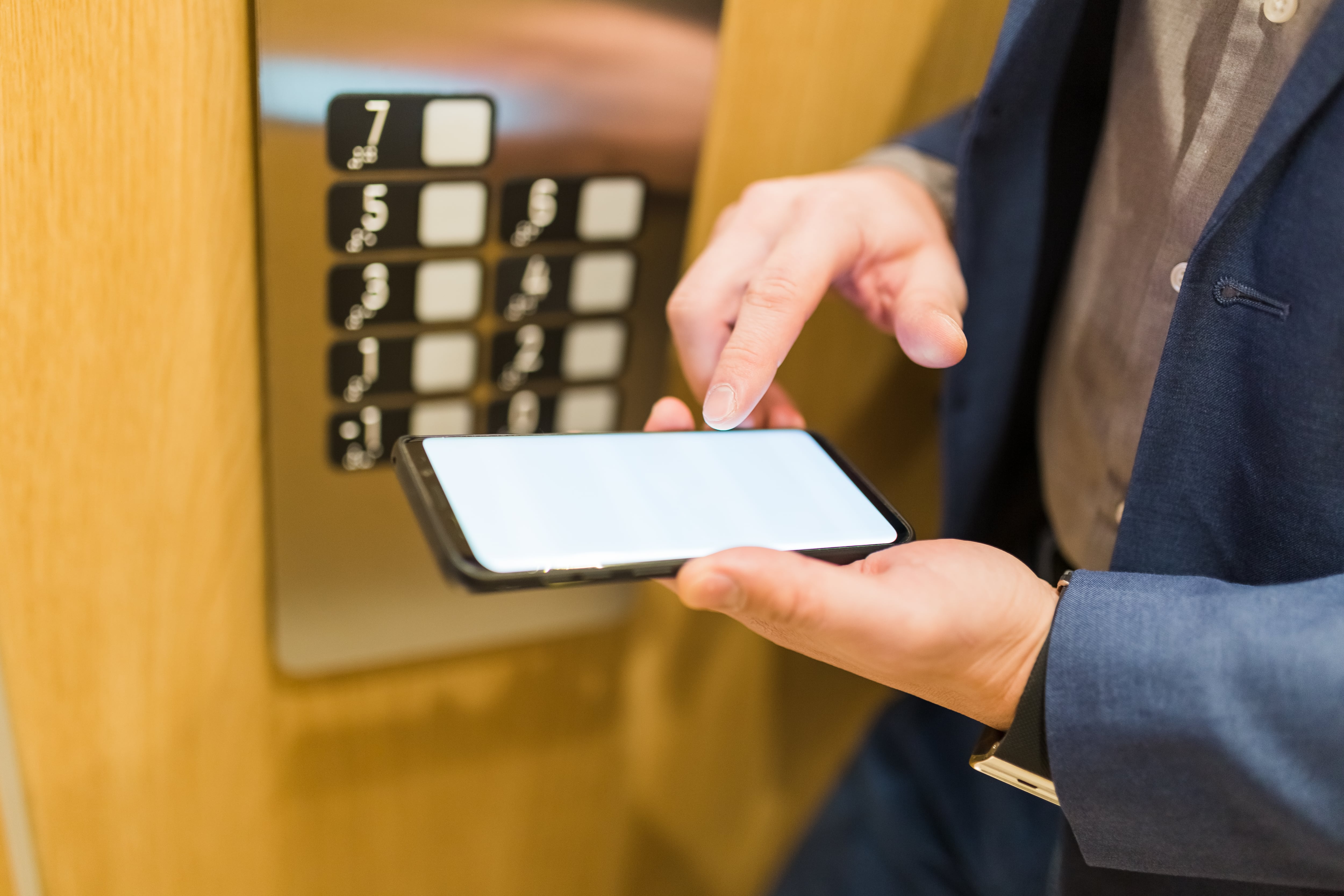 Close up of businessman using blank screen smartphone next to elevator control panel. Business and office building meeting concept.