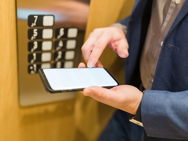 Close up of businessman using blank screen smartphone next to elevator control panel. Business and office building meeting concept.