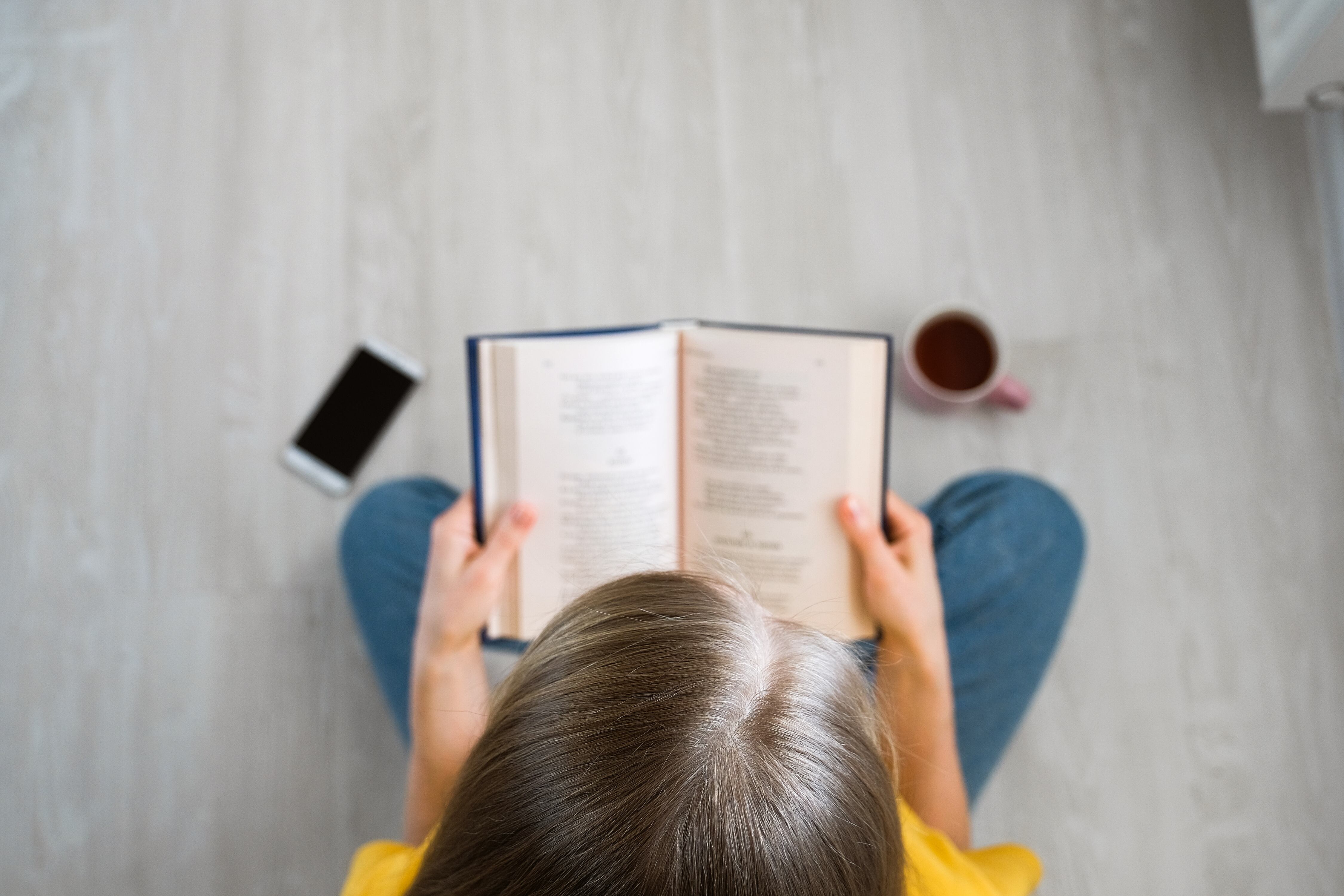 The girl is sitting on the floor with her legs crossed in the lotus position and reading a book. A woman holds a textbook in her hands. Next to it is a mug of tea or coffee and a mobile phone. The concept of education, development and self-improvement.