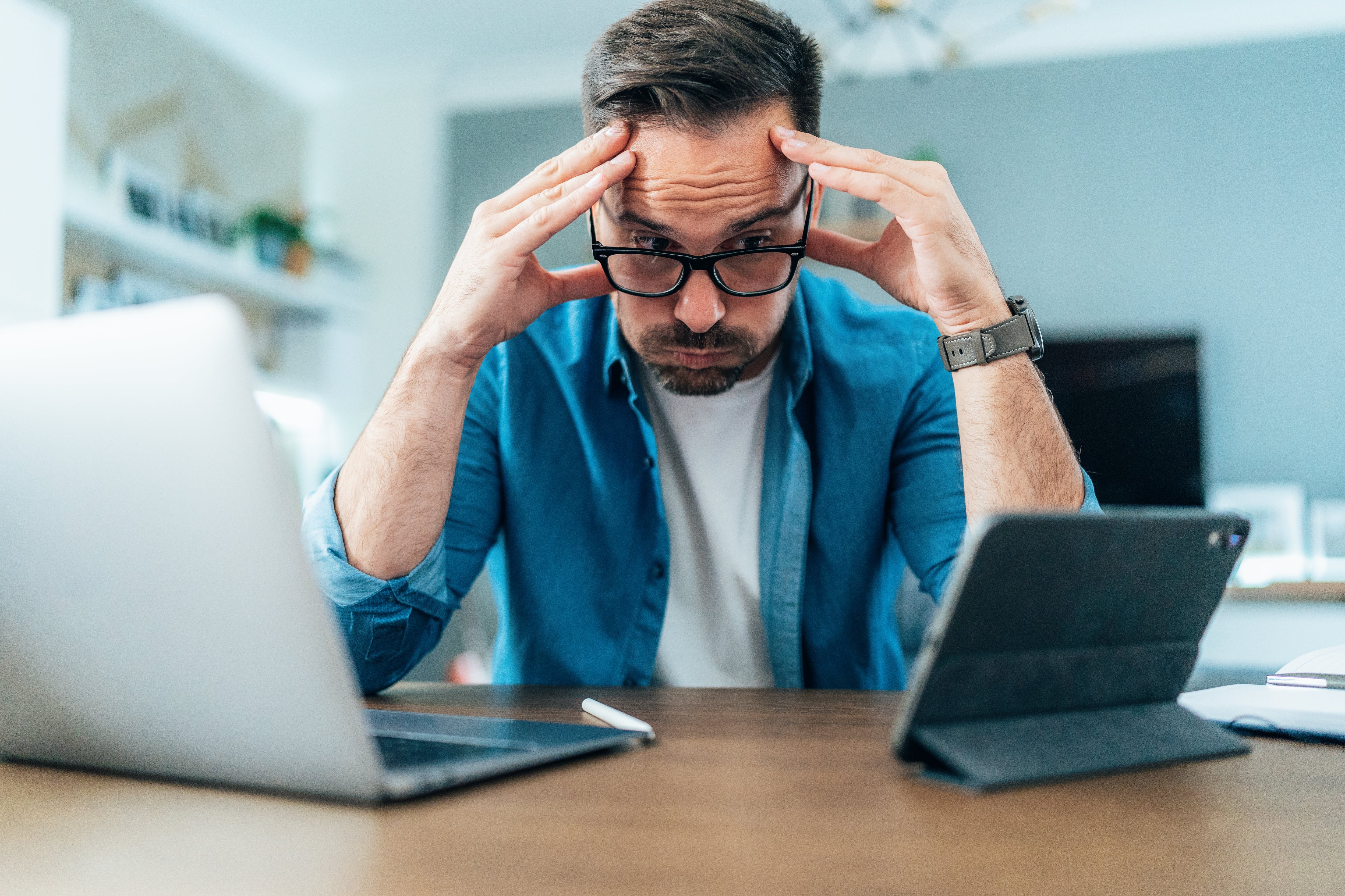 Tired Businessman working at home using laptop and digital tablet. Overworked young man with eyeglasses looking at laptop bored and anxious. Man with headache holding head in hands in front of digital devices.
