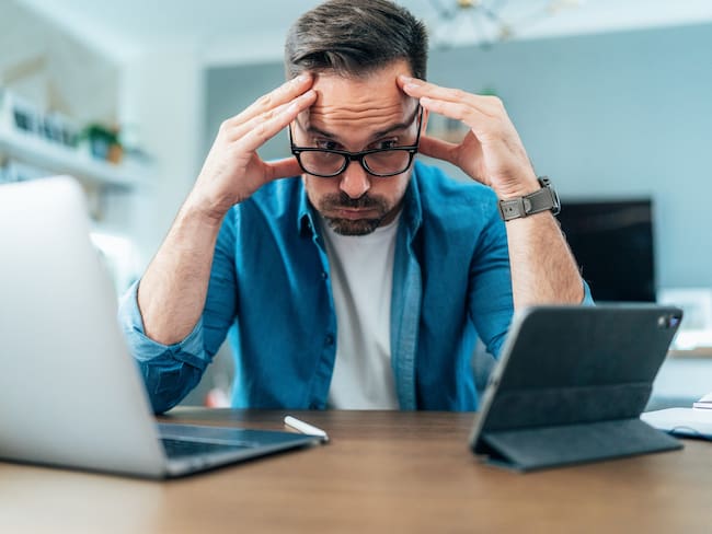Tired Businessman working at home using laptop and digital tablet. Overworked young man with eyeglasses looking at laptop bored and anxious. Man with headache holding head in hands in front of digital devices.