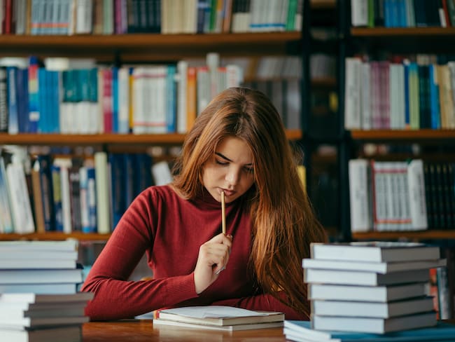 Mujer sentada en la mesa de una biblioteca leyendo unos apuntes mientras sostiene un lápiz (Getty Images)