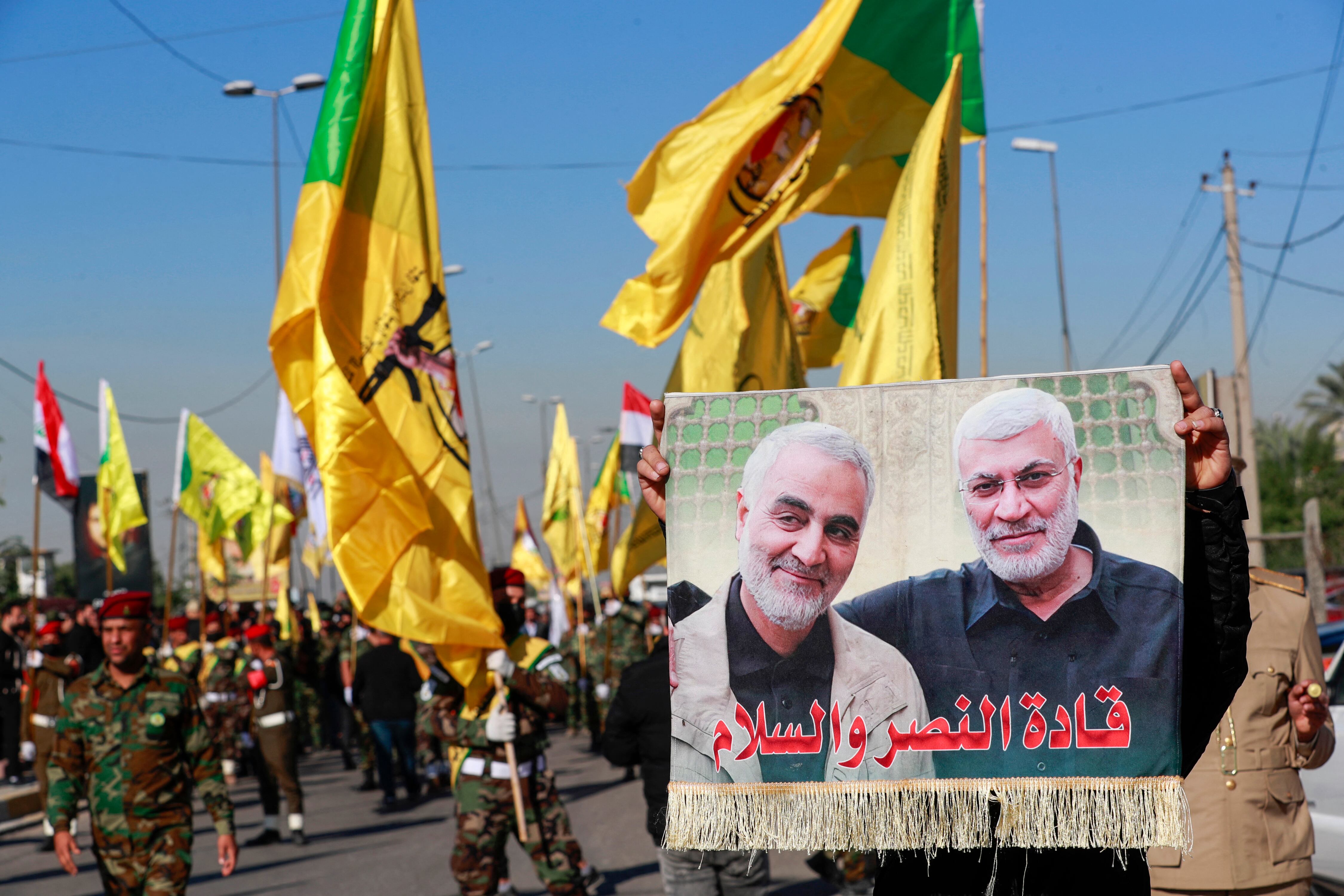 A man lifts a placard depicting slain Iranian commander Qasem Soleimani (L) and Iraqi commander Abu Mahdi al-Muhandis, as fighters bearing flags of Iraq and paramilitary groups, including al-Nujaba and Kataib Hezbollah, march during a funeral in Baghdad for five militants killed a day earlier in a US strike in northern Iraq, on December 4, 2023. Dozens of mourners gathered on December 4 for the funeral of the five militants killed the previous day near Kirkuk in what a US military official described as a "self-defense strike". (Photo by AHMAD AL-RUBAYE / AFP) (Photo by AHMAD AL-RUBAYE/AFP via Getty Images)