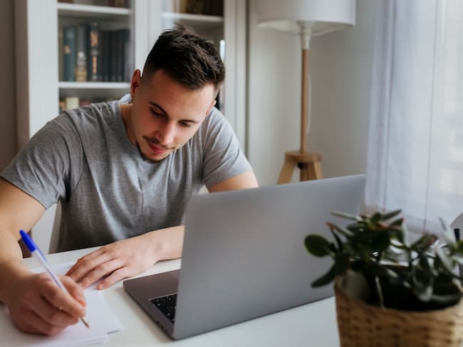 Hombre escribiendo en papel mientras está sentado con un computador portátil en su casa / Foto: GettyImages
