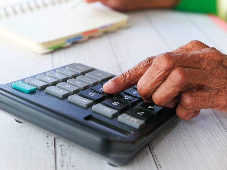 Hombre mayor haciendo cálculos en calculadora con un cuaderno en el fondo (Getty Images)