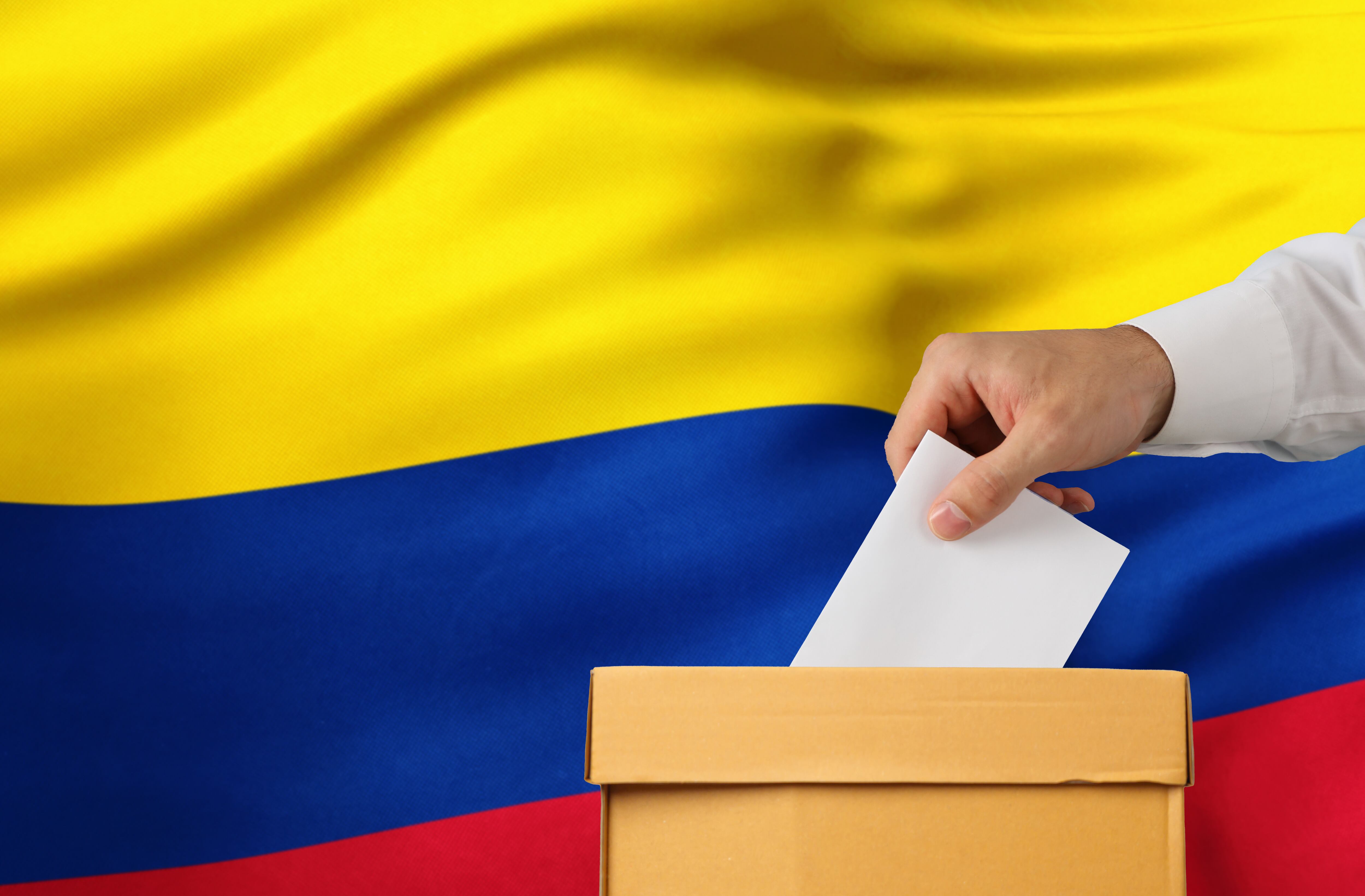 Deposito de voto en la urna electoral y bandera de Colombia al fondo - Primer plano.