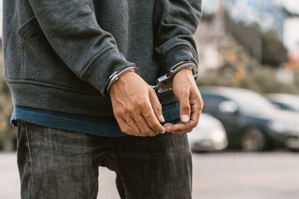 Under arrest, a man's hands with clenched fists are handcuffed behind him. Black background with copy space.