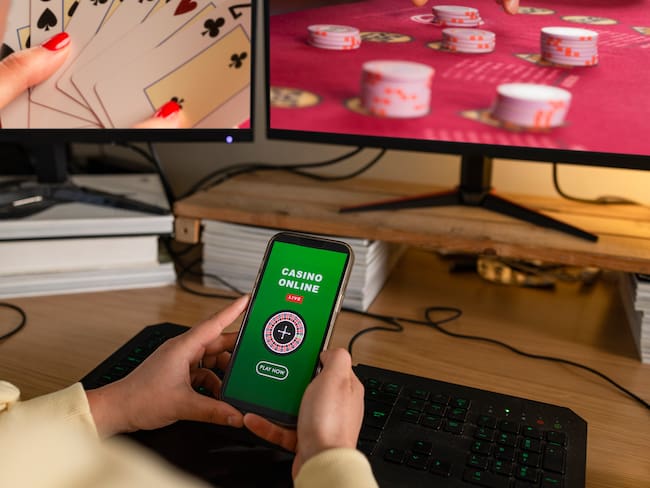 A rear-view shot of an unrecognisable young woman sitting at a desk with online gambling on her computer screens, she has a mouse and keyboard. She is also using her smart phone to gamble in an online casino.