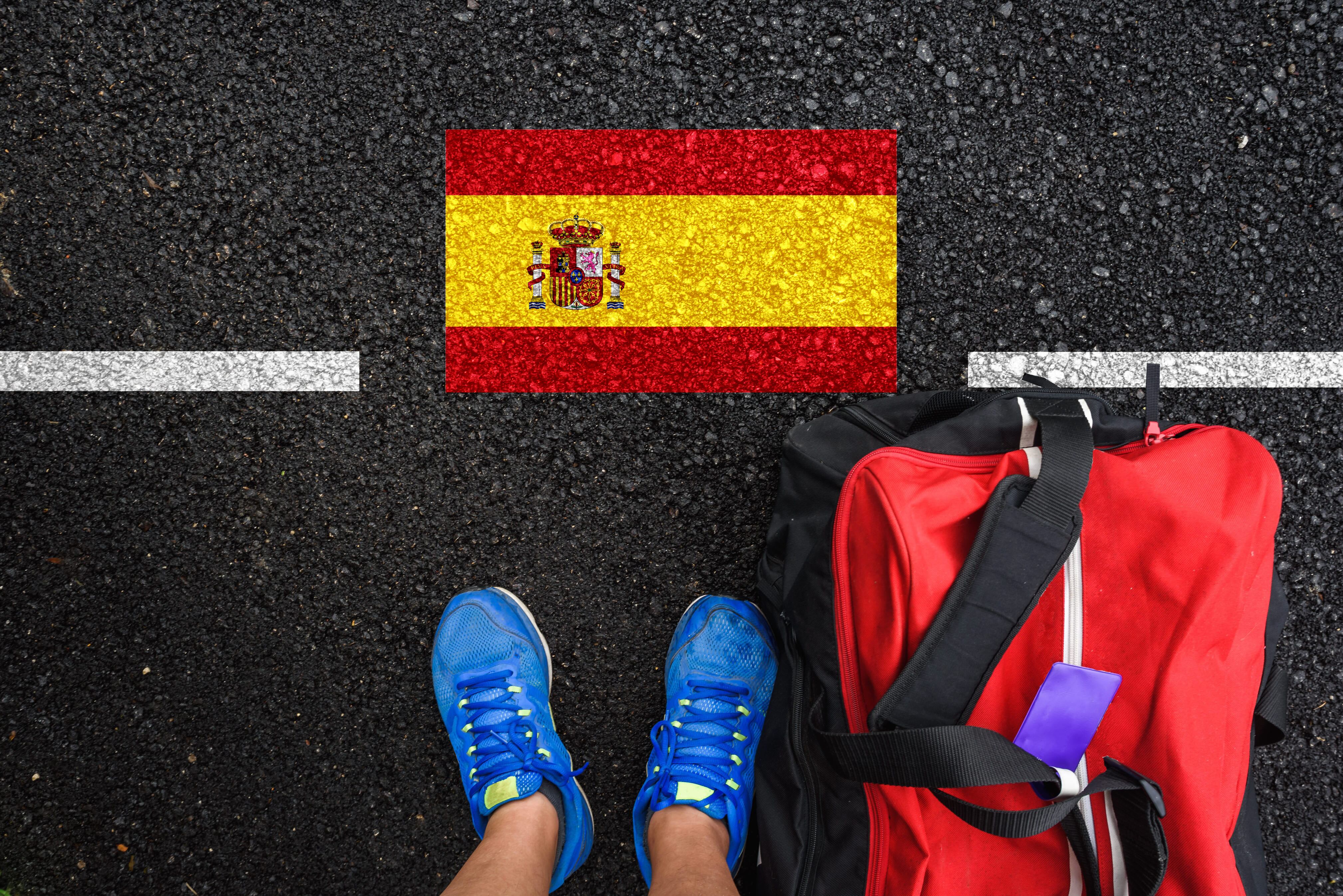 En la imagen se ve a un hombre junto con una maleta de viaje, parado enfrente de la bandera de España / Foto: GettyImages