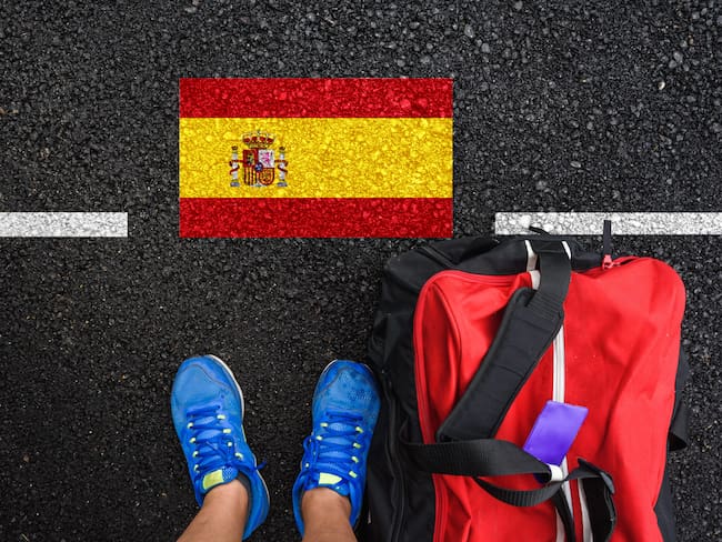 En la imagen se ve a un hombre junto con una maleta de viaje, parado enfrente de la bandera de España / Foto: GettyImages