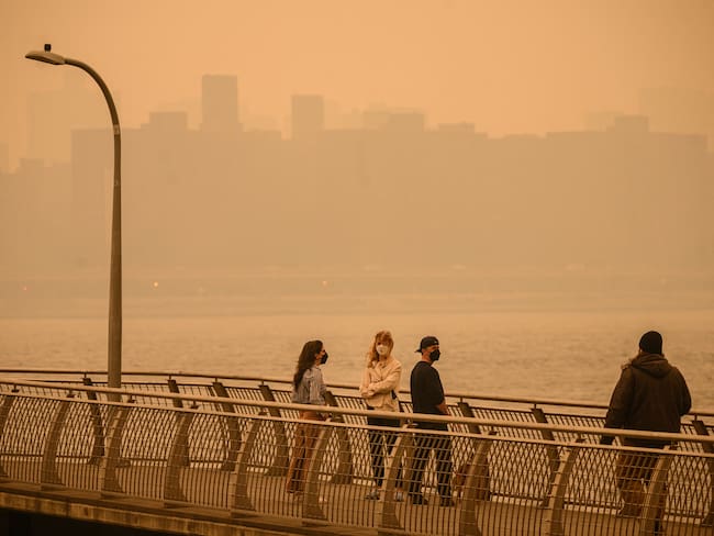 The Manhattan skyline is seen past people wearing face masks while walking along a pier as smoke from wildfires in Canada cause hazy conditions in New York City on June 7, 2023. An orange-tinged smog caused by Canada&#039;s wildfires shrouded New York on Wednesday, obscuring its famous skyscrapers and causing residents to don face masks, as cities along the US East Coast issued air quality alerts. (Photo by Ed JONES / AFP)