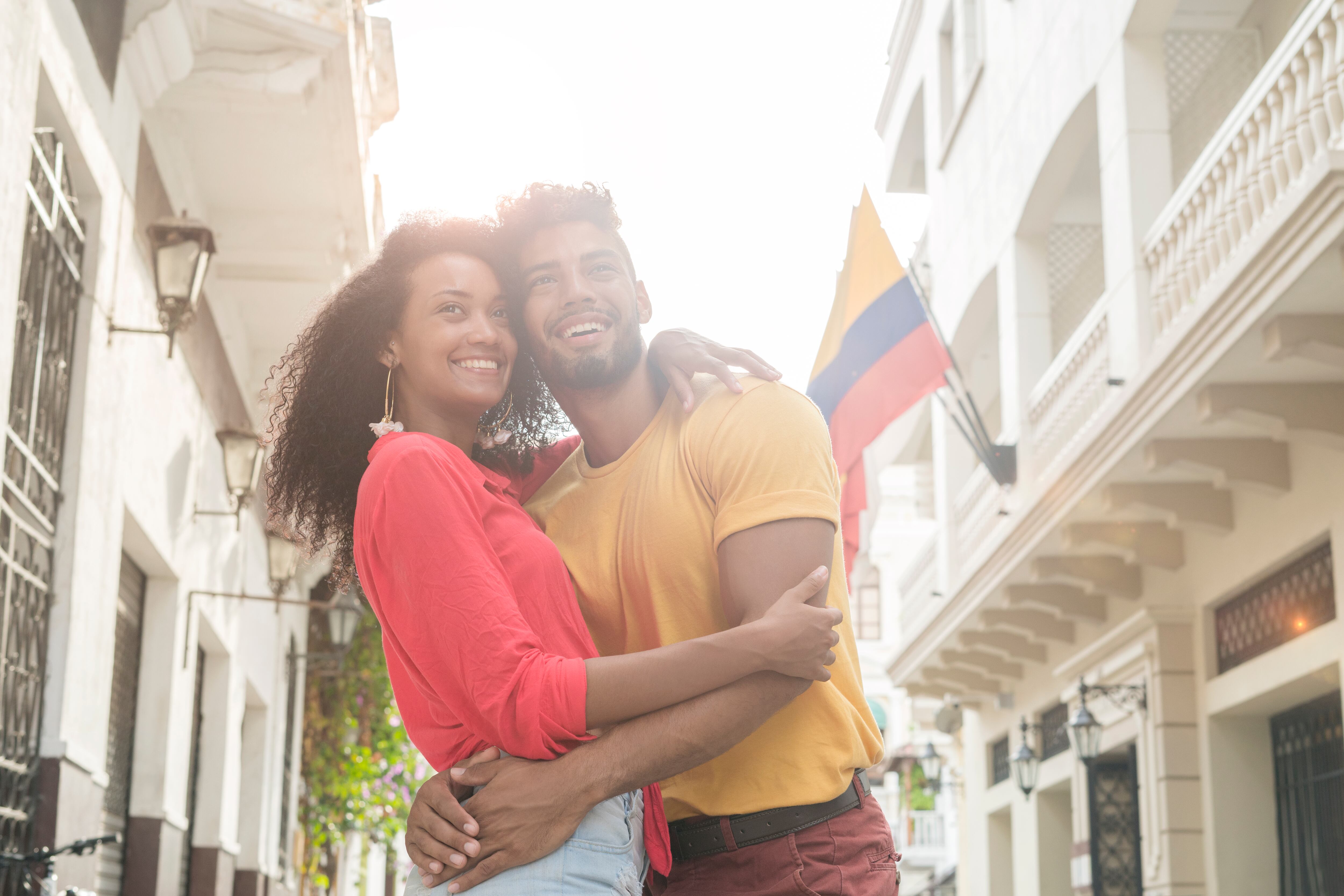 Latin couple made up of black curly-haired woman in brown skin, red blouse and jean short that is hugging her boyfriend Latin man with curly hair and yellow sweater who is also hugging her while they know the historic center of the city of Cartagena where We observe old house with flags of Colombia