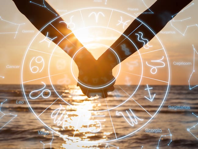 De fondo, una pareja sosteniendo sus manos frente a la playa. Encima, la rueda del zodiaco (Foto vía GettyImages)