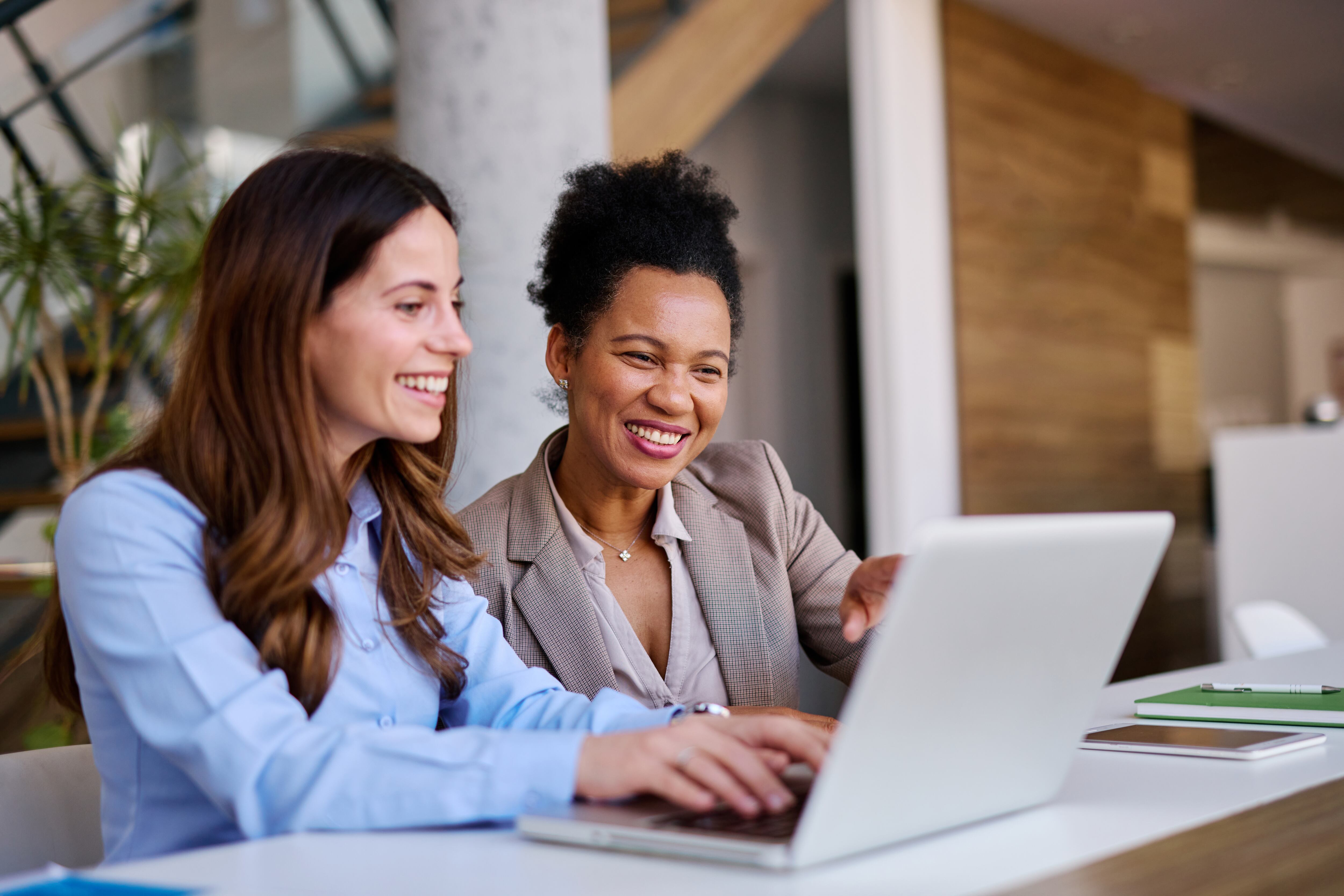 Two businesswomen are collaborating on a project, looking at a laptop and smiling, demonstrating teamwork and productivity in a modern office environment