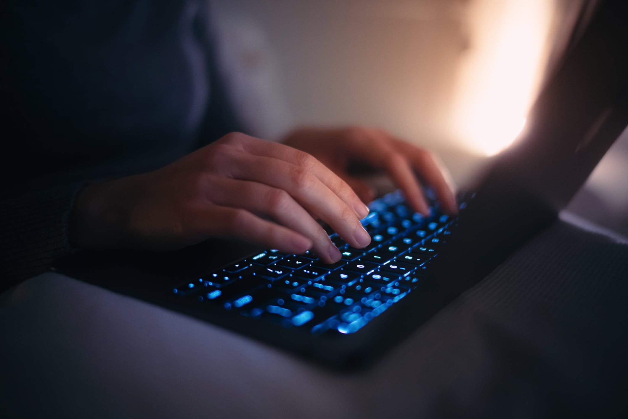 Close up hands of female using laptop in bed at night. Woman's hand typing on keyboard, working late, device screen light illuminated on her. Lifestyle and technology