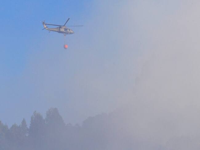 BOG400. BOGOTÁ (COLOMBIA), 22/01/2024.- Un helicóptero, que utiliza el sistema Bambi bucket, arroja agua entre el humo que se observa hoy en los cerros orientales en Bogotá (Colombia). Bomberos, la Defensa Civil y las fuerzas de seguridad trabajan desde la mañana de este lunes para extinguir un incendio en los cerros de Bogotá que ha llenado de humo el oriente de la capital colombiana. EFE/ Carlos Ortega
