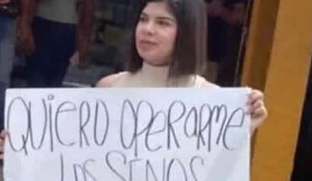 Una mujer que sostiene un aviso con un polémico mensaje que dice "quiero operarme los senos, ¿me ayudas?",