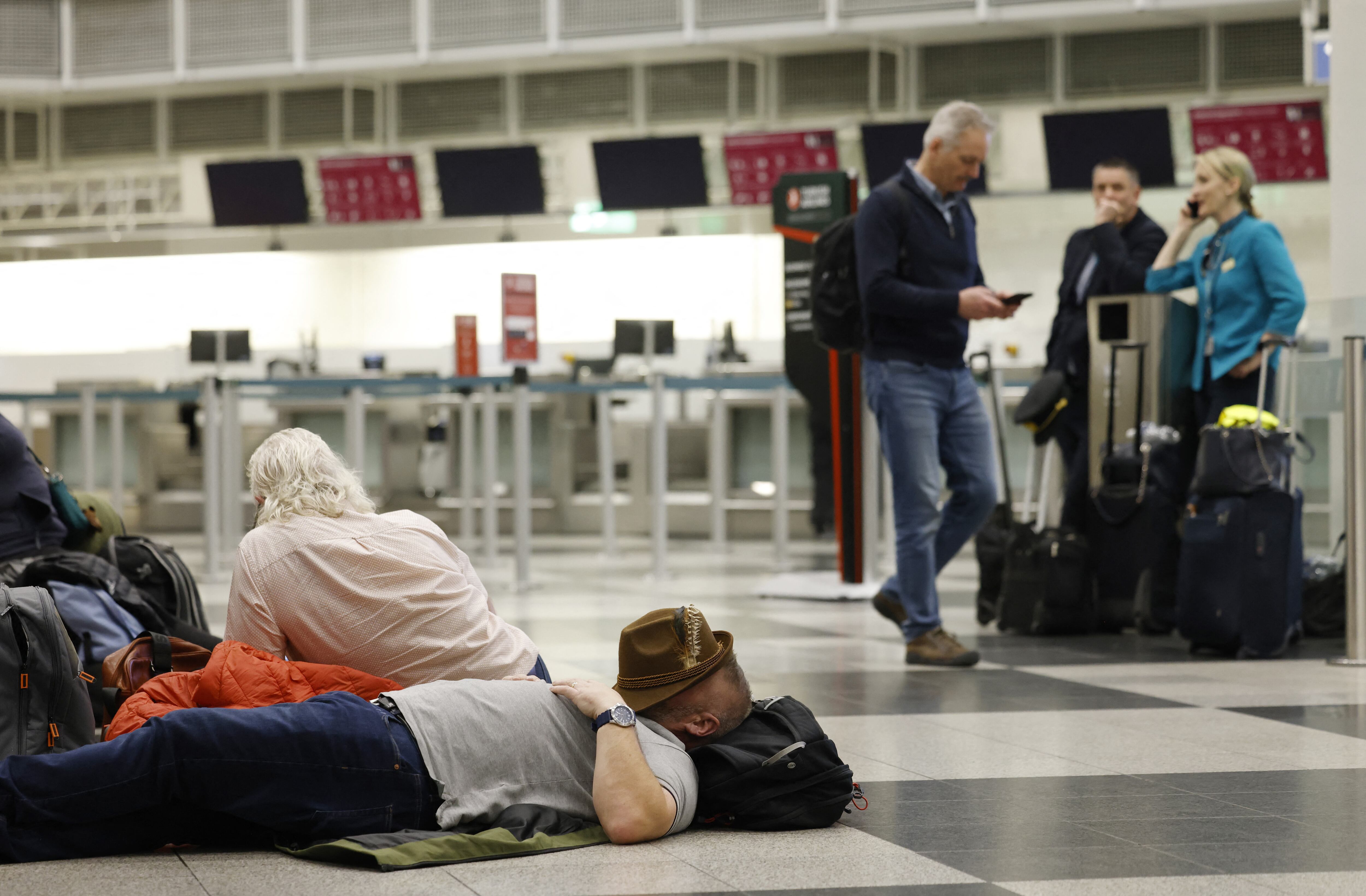 A passenger has his covered his face with a hat to sleep on the floor at Munich International Airport in Munich, southern Germany, on early October 4, 2025, after reports of drone sightings again forced Munich airport to close and more than 30 flights had to be cancelled and left nearly 3,000 passengers stranded. New reports of drone sightings again forced Munich airport to close late on October 3, 2025, a day after drones caused more than 30 flights to be cancelled there and left nearly 3,000 passengers stranded. The airport said in a statement on its website that "because of unconfirmed drone sightings air traffic has been cancelled until further notice", the latest in a spate of such incidents heightening security fears in Europe. (Photo by Michaela STACHE / AFP)