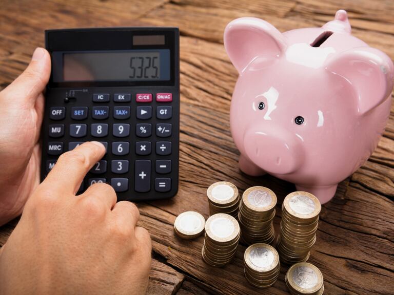 Hombre usando la calculadora, junto con una alcancía de monedas ubicada sobre una mesa de madera (Getty Images)