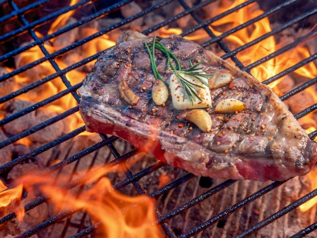 Filete ribeye a la parrilla en una parrilla de carbón caliente con llamas cubiertas con dientes de ajo salteados una palmadita de mantequilla y romero // Getty images